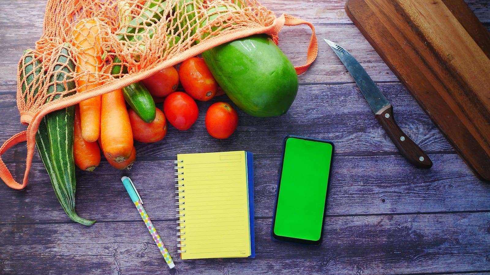 Verdure in una retina, un blocco per appunti giallo, una penna e uno smartphone su un tavolo di legno