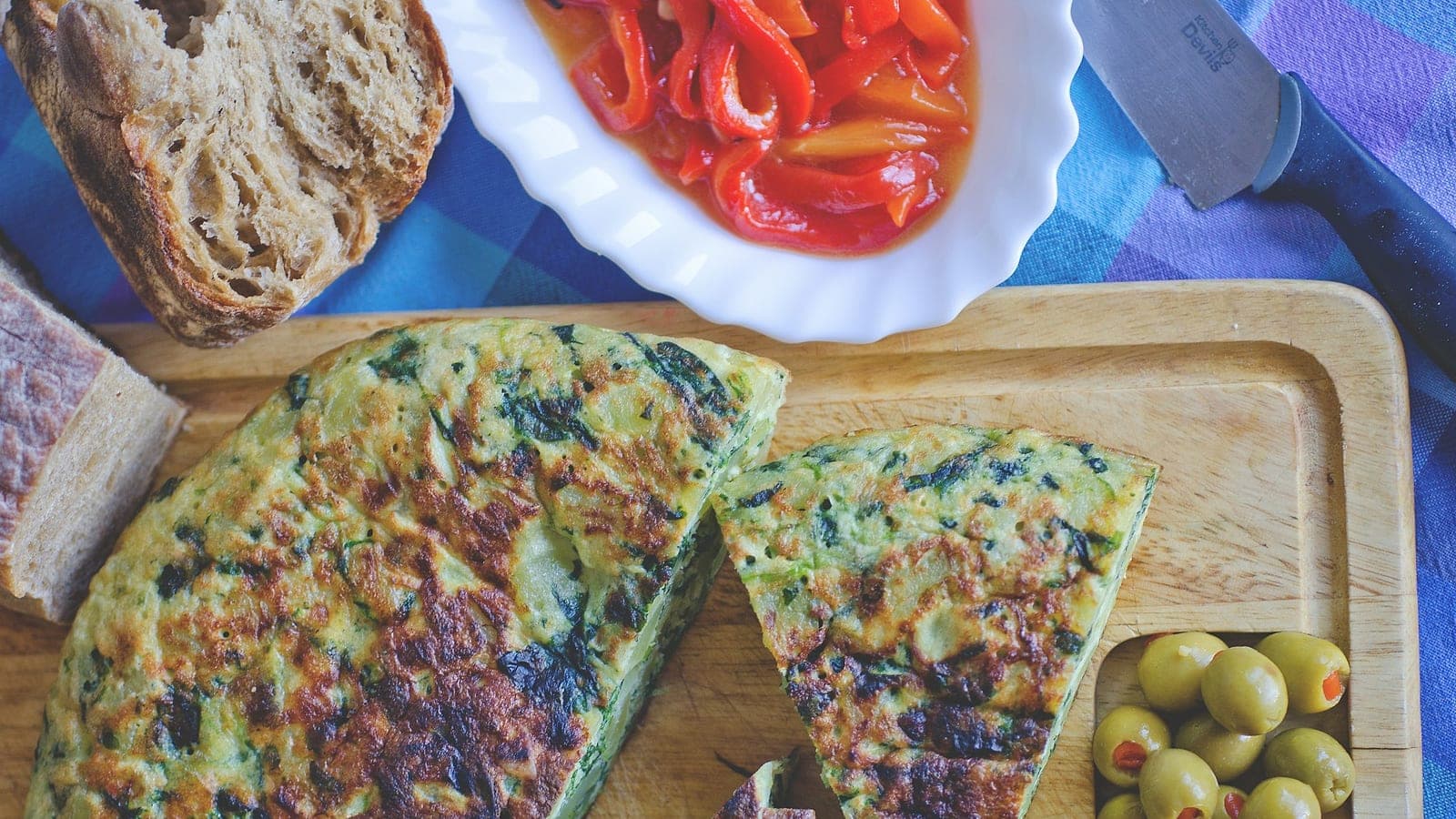 Una frittata di spinaci e patate tagliata su un tagliere, con olive e peperoni arrostiti