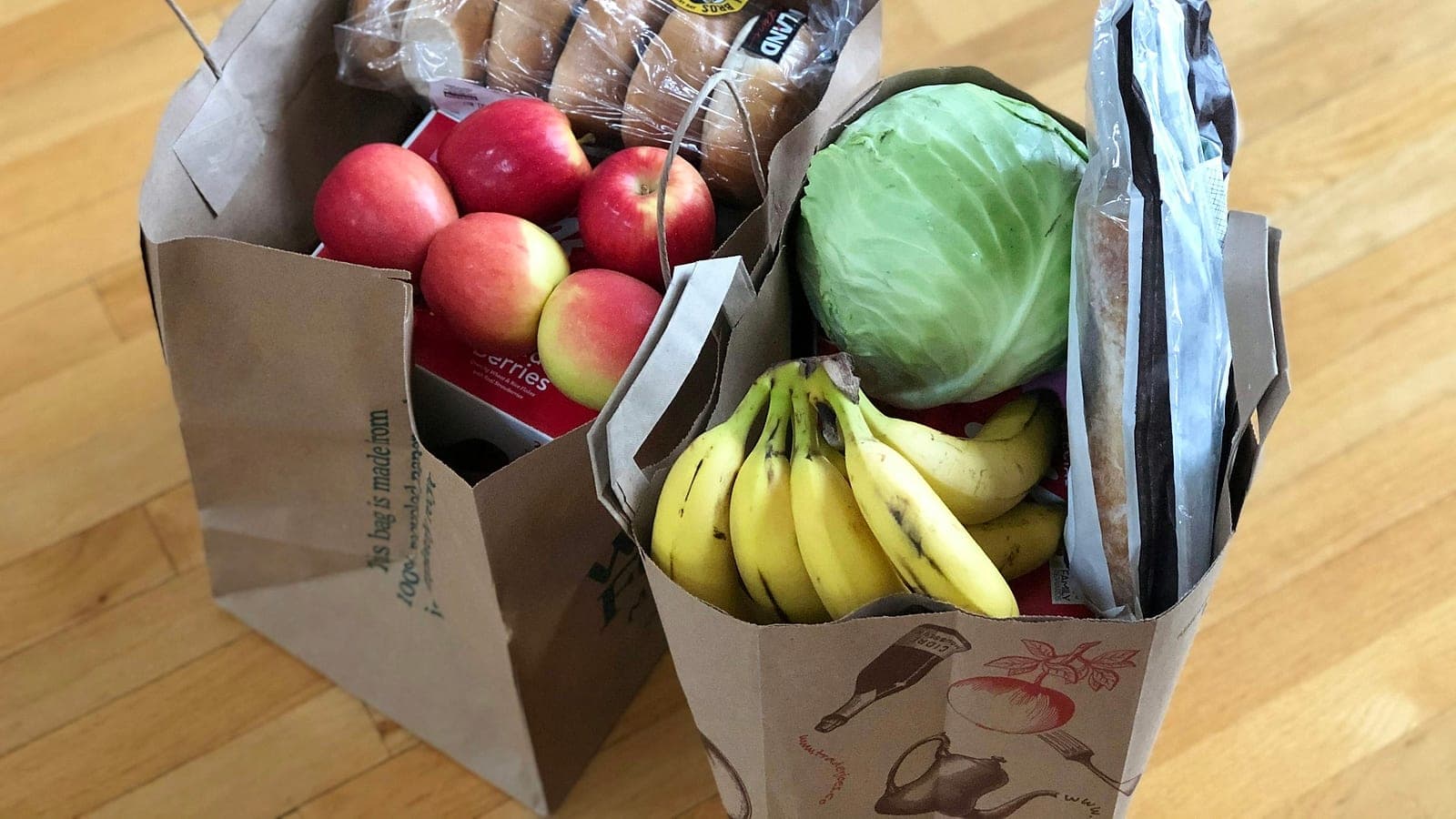 Due sacchetti di carta della spesa con mele, banane, un cavolo e pane, appoggiati su un pavimento di legno